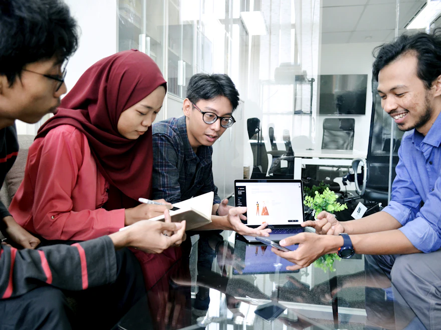 Seorang wanita bertudung dan tiga rakan sepasukan berbincang di sekeliling komputer riba
