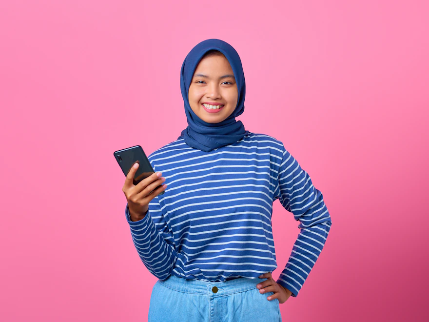 Seorang wanita bertudung tersenyum sambil memegang telefon bimbit