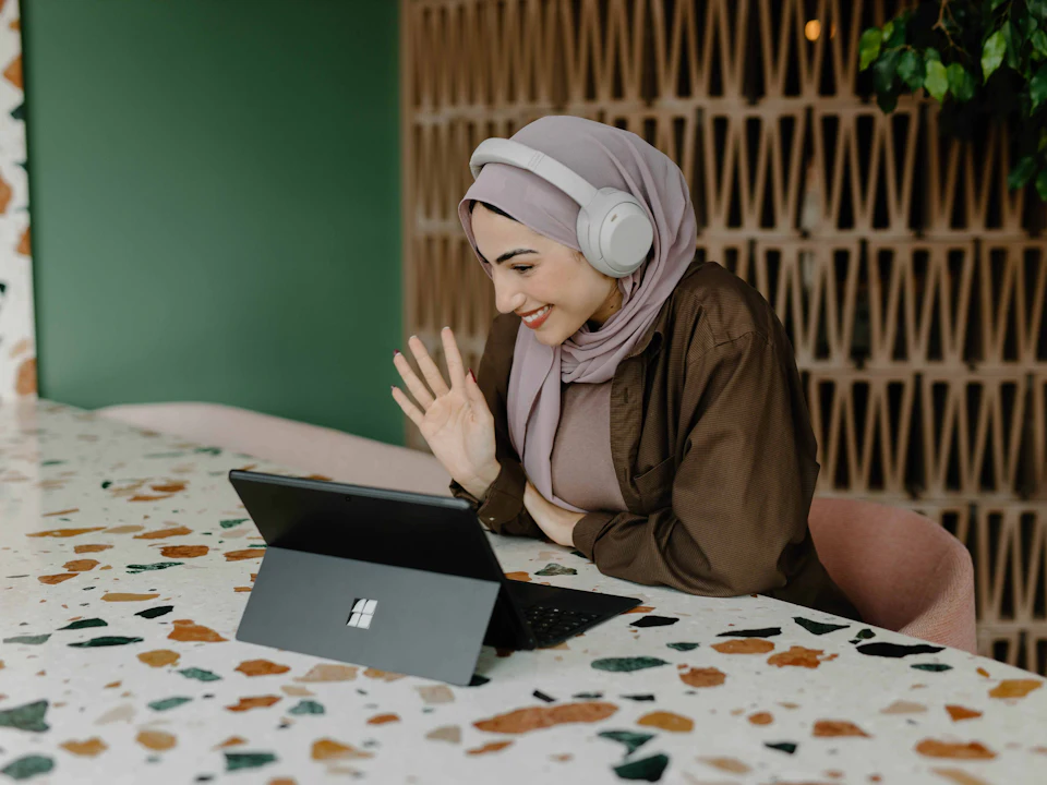 Seorang wanita bertudung memakai fon kepala melambai semasa panggilan video di komputer riba
